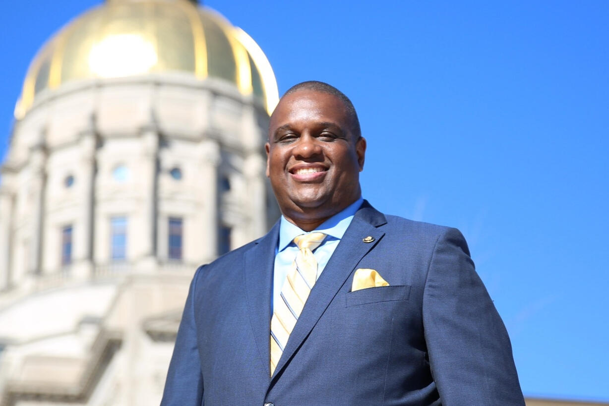 Otha is pictured in a navy blue suit and yellow striped tie. He is smiling in front of the capital building.